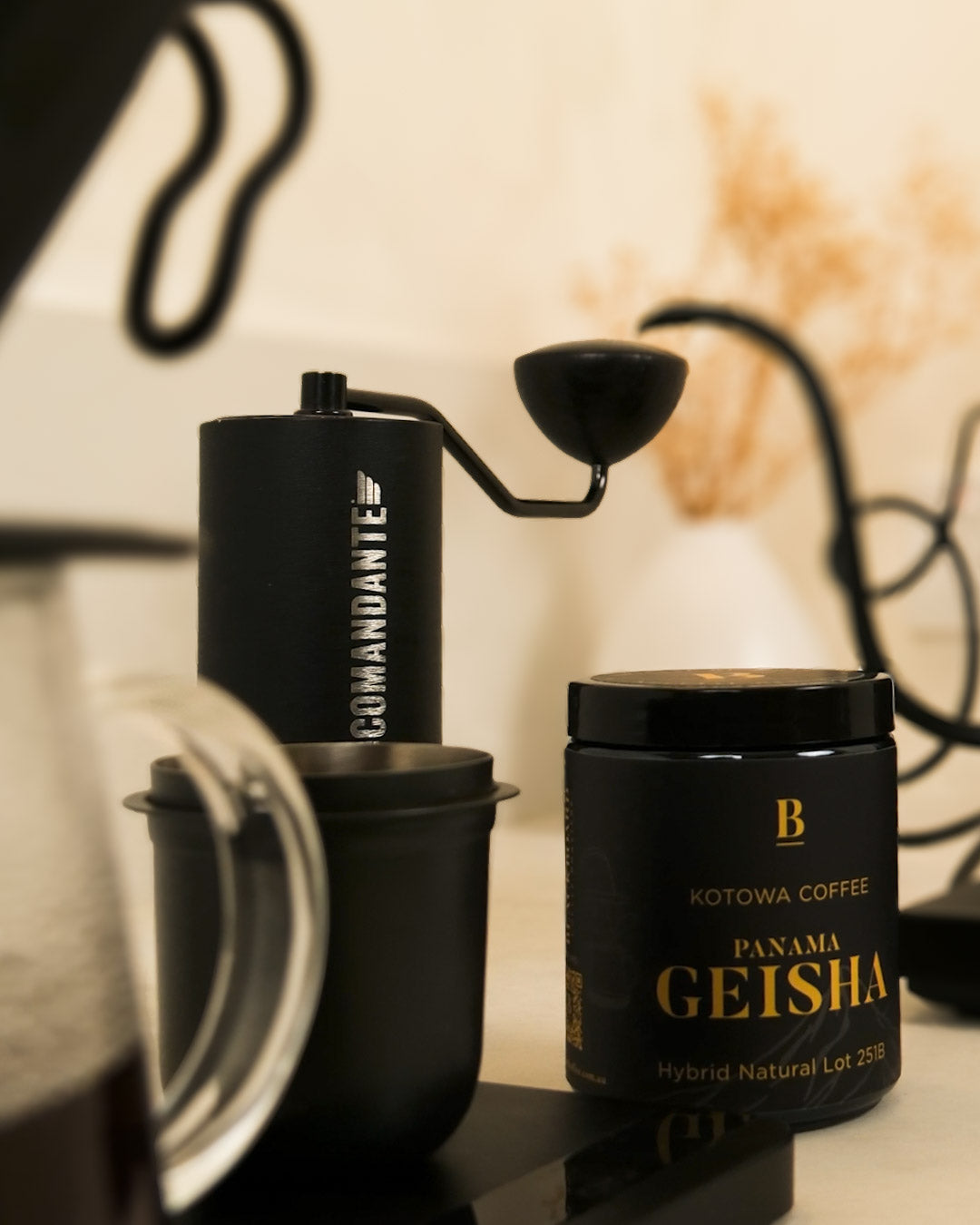 Black coffee grinder and Kotowa Coffee Panama Geisha canister on a reflective surface with a blurred background.