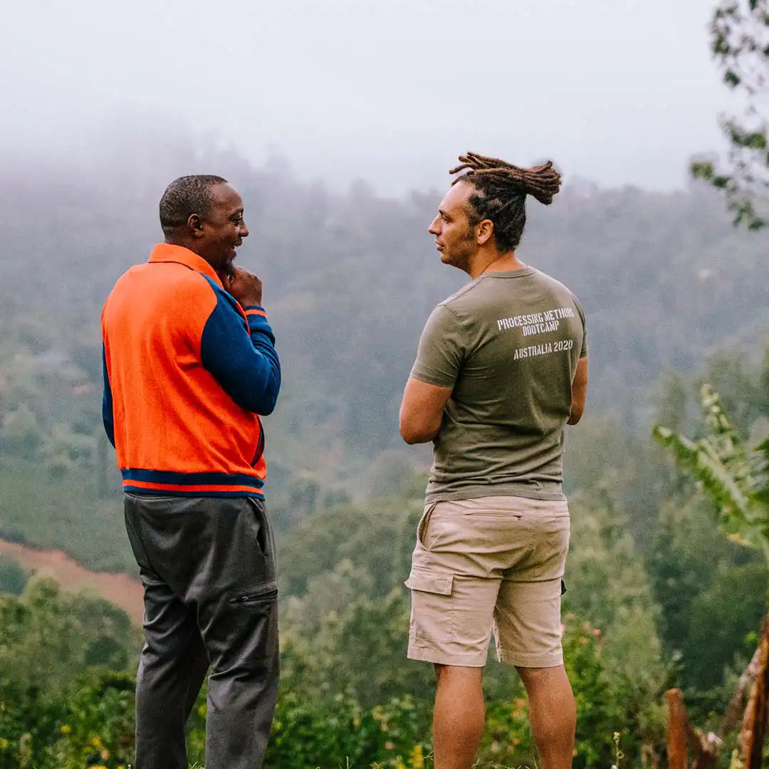 Two men standing outdoors in a misty forested coffee  landscape, wearing Process of Destruction apparel. One man wears an orange and navy sweater vest over a blue shirt, and the other wears an olive green t-shirtshorts.